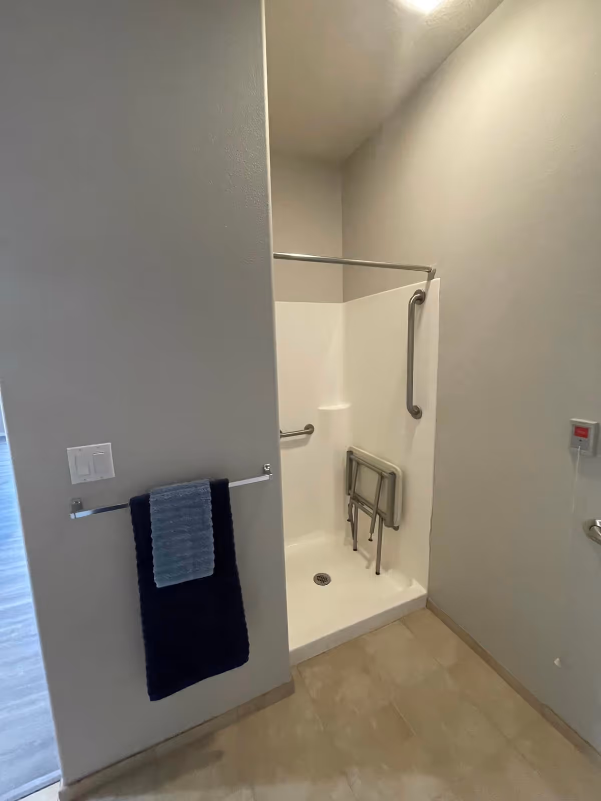 A bathroom shower area with a foldable shower seat and grab bars for accessibility. There is a towel rack on the wall outside the shower holding two towels, one dark blue and one light blue. The floor is tiled and the walls are painted light gray.