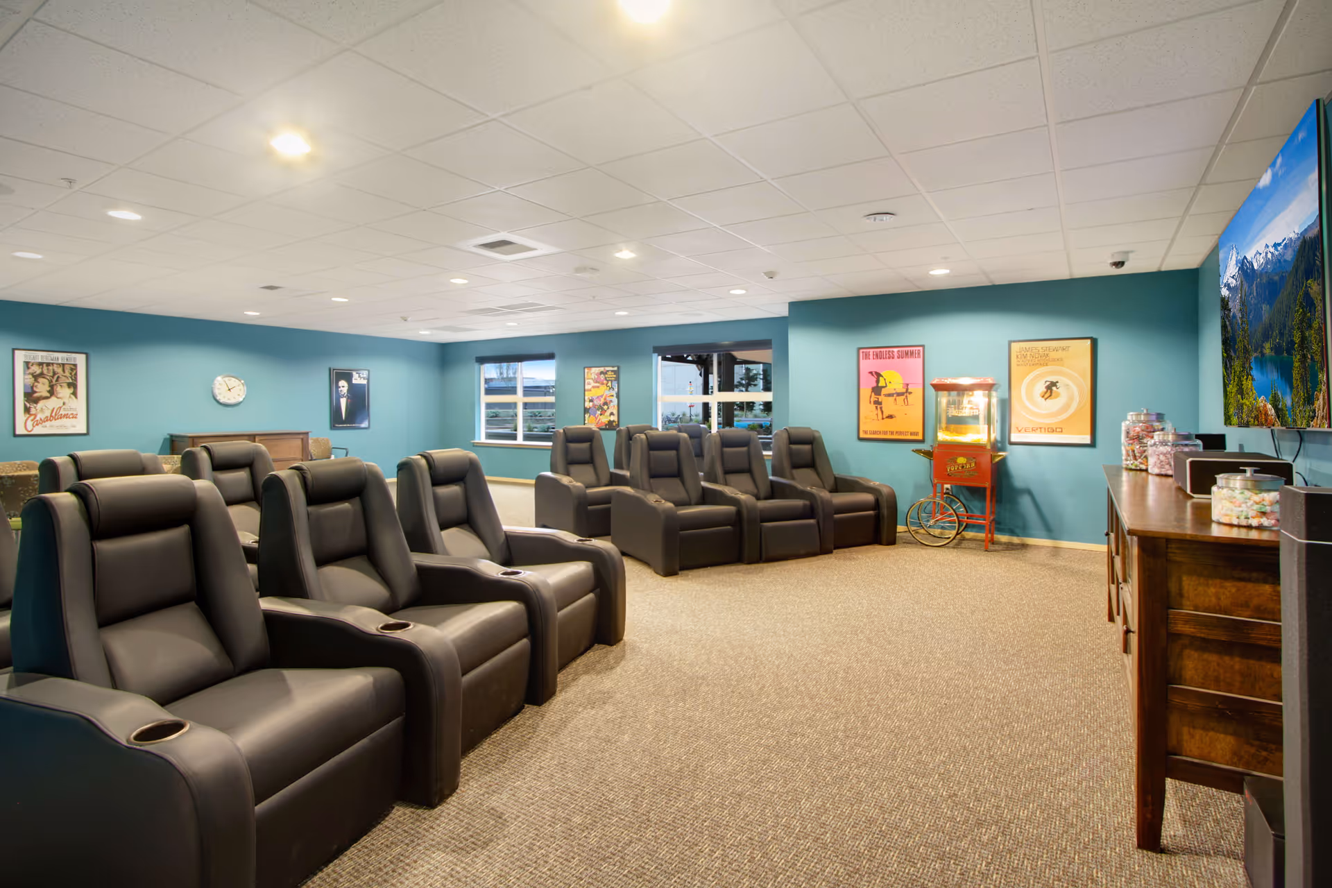 A lounge-style media room with rows of black reclining theater chairs, teal walls, wall art, a popcorn machine and a large TV.