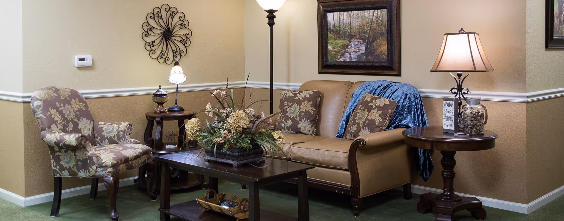 A cozy living room area with a tan leather sofa adorned with floral pillows and a blue throw blanket. Next to the sofa is a round wooden side table with a lamp, a jar, and a small framed sign. There is a floral upholstered armchair to the left, a wooden coffee table with a floral centerpiece, and a floor lamp behind the sofa. A framed painting of a forest scene hangs on the beige wall.
