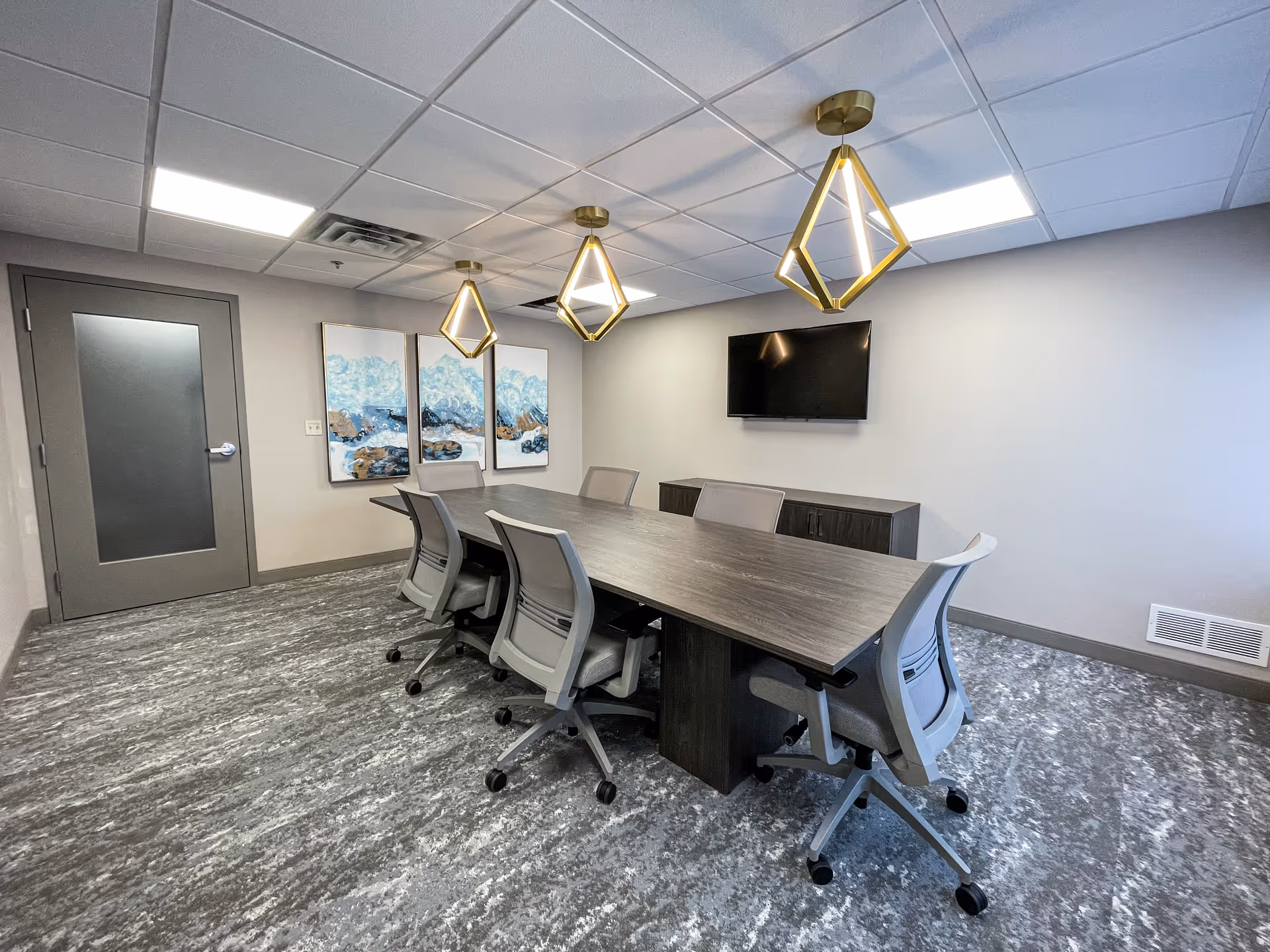 A modern conference room with a long rectangular table surrounded by six office chairs on wheels. Three geometric pendant lights hang from the ceiling above the table. On the wall opposite the door, there is a flat-screen TV mounted above a low cabinet. Three abstract blue and brown paintings are displayed on the wall to the left of the TV. The room has a gray door, gray carpet with a textured pattern, and a white ceiling with recessed lighting.