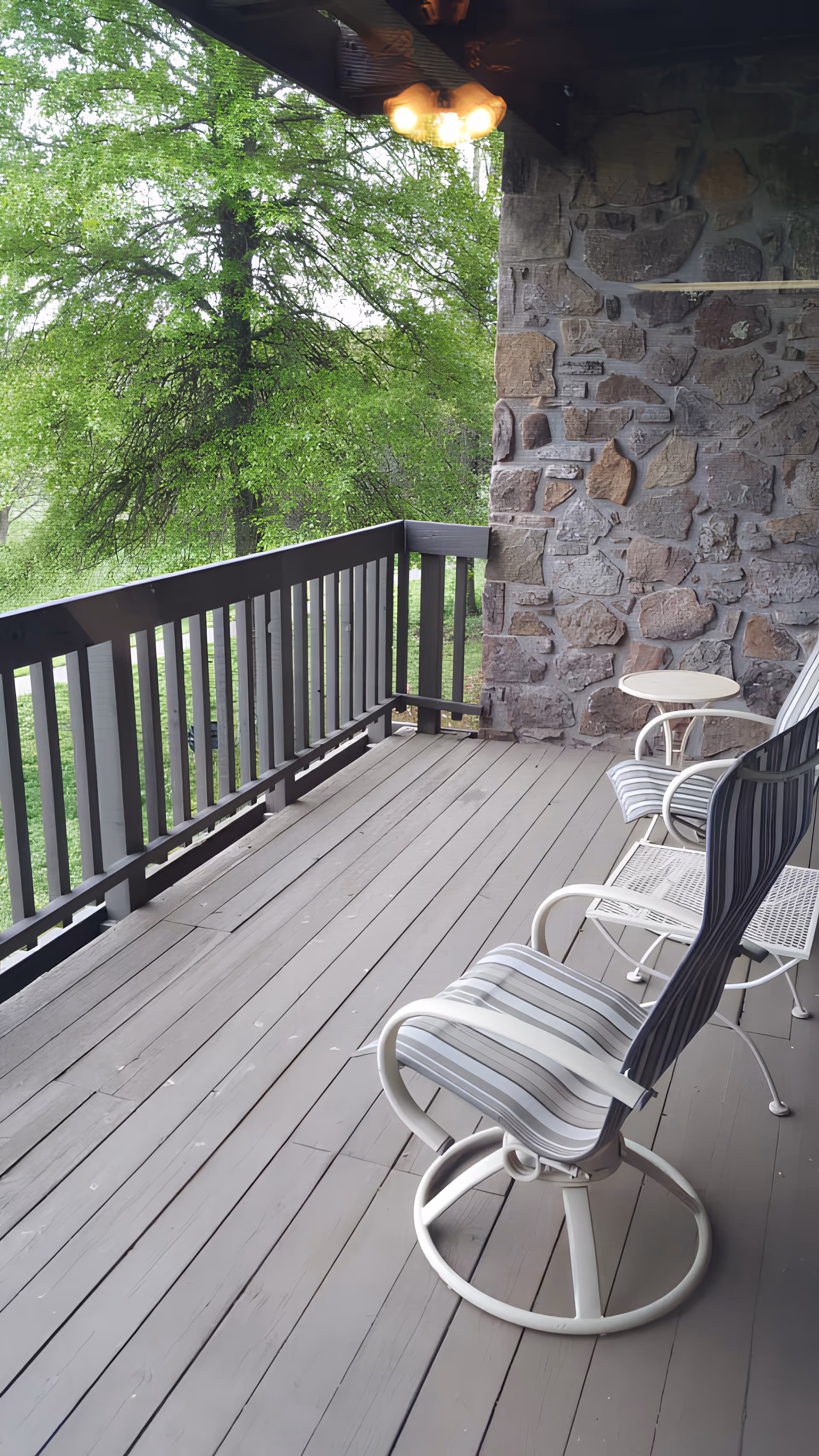 Covered wooden balcony with striped patio chairs and a small side table next to a stone wall overlooking green trees.