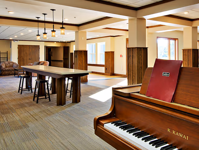 A bright communal lounge with a piano in the foreground, a long table with stools, and seating areas by large windows.