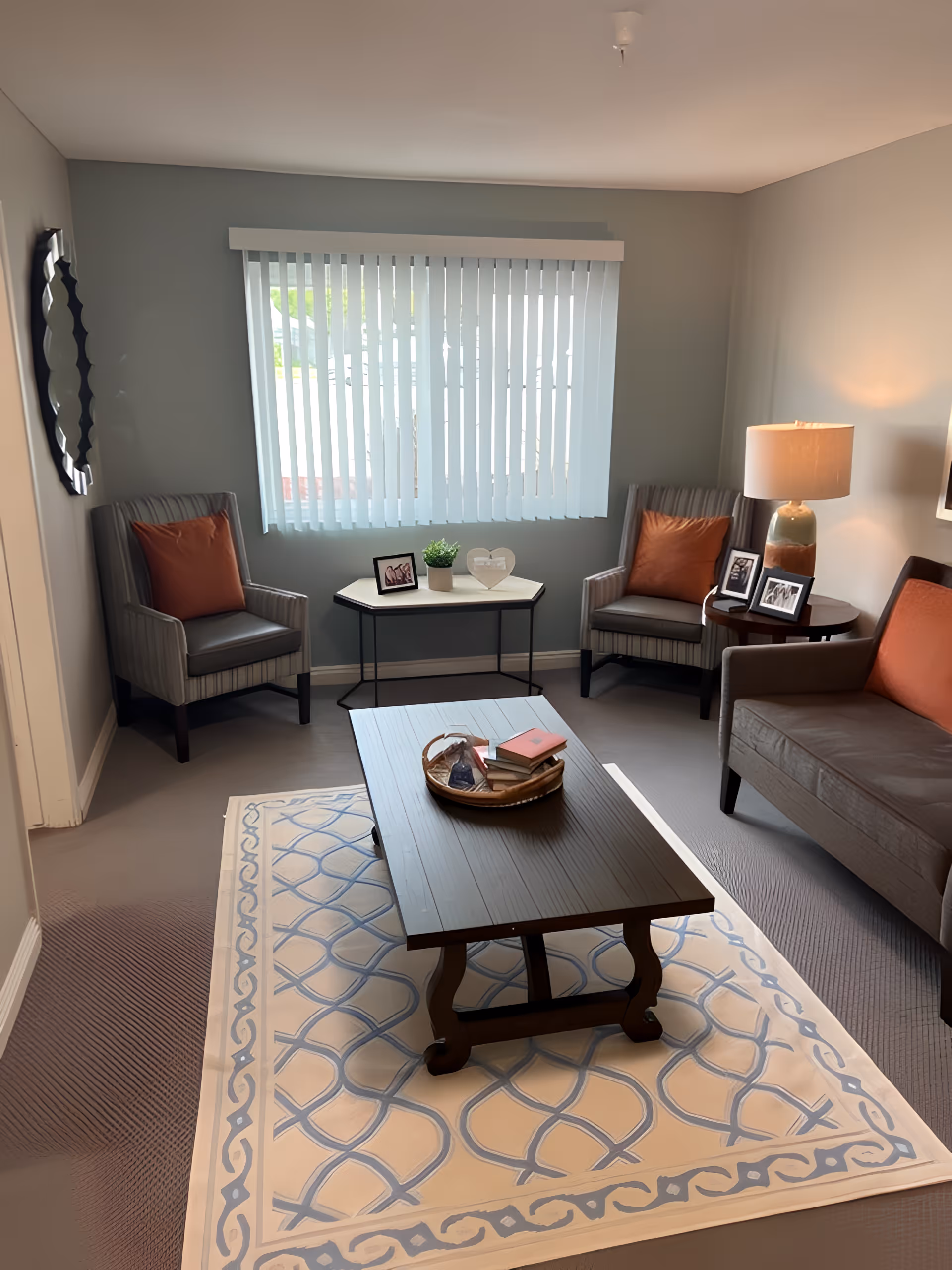 A cozy living room with a gray sofa and two striped armchairs, each with an orange cushion. A wooden coffee table sits on a patterned rug in the center, holding a tray with books and decorative items. A small table with a lamp and framed photos is next to the sofa, and a window with vertical blinds lets in natural light.