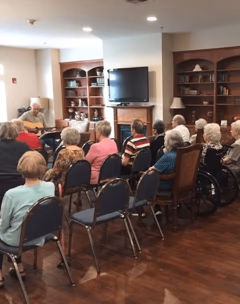 A group of elderly people seated in chairs facing a man playing a guitar in a cozy room with wooden bookshelves, a fireplace, and a mounted flat-screen TV.