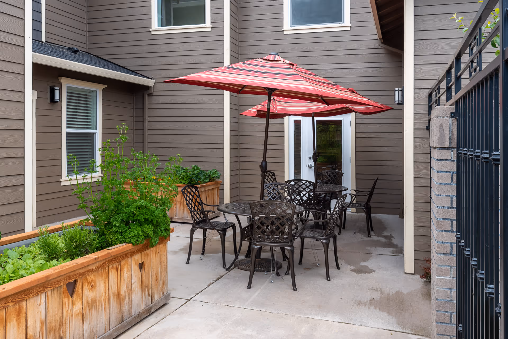 Outdoor patio area with a round metal table and six matching chairs under a large red and white striped umbrella. There are two wooden planter boxes with green plants along the left side, and the patio is surrounded by brown siding walls with windows and a glass door. A black metal fence is visible on the right side.