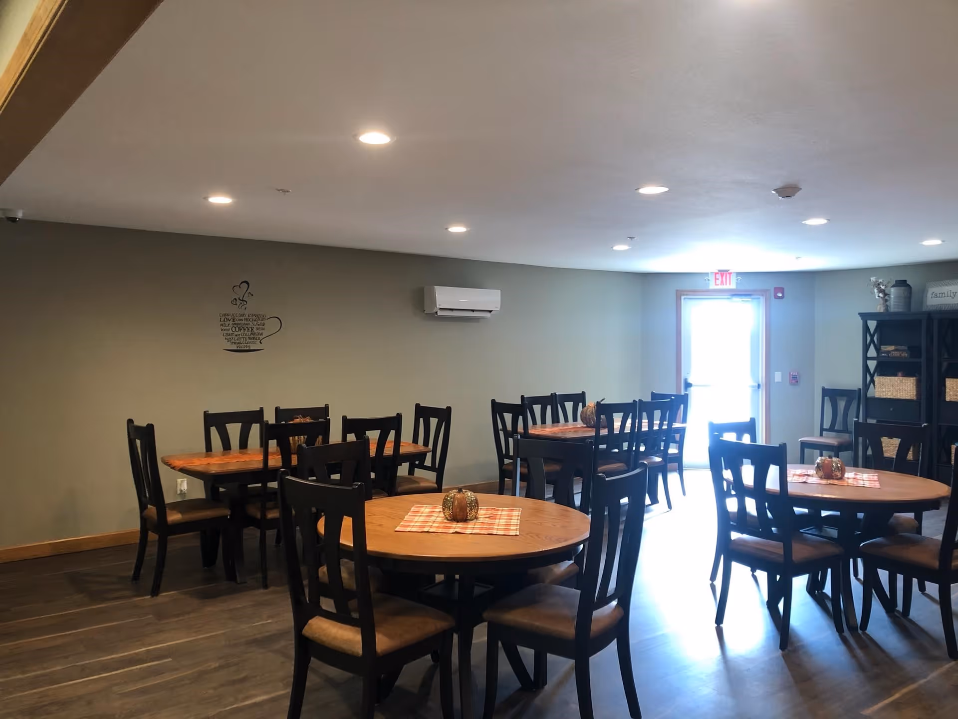 A dining room with several wooden tables and chairs arranged neatly. Each table has a small decorative centerpiece on a checkered placemat. The room has a light gray wall with a coffee cup wall decal, recessed ceiling lights, a wall-mounted air conditioning unit, and a door with an exit sign above it. There is also a black shelving unit with baskets and decor on the right side of the room.