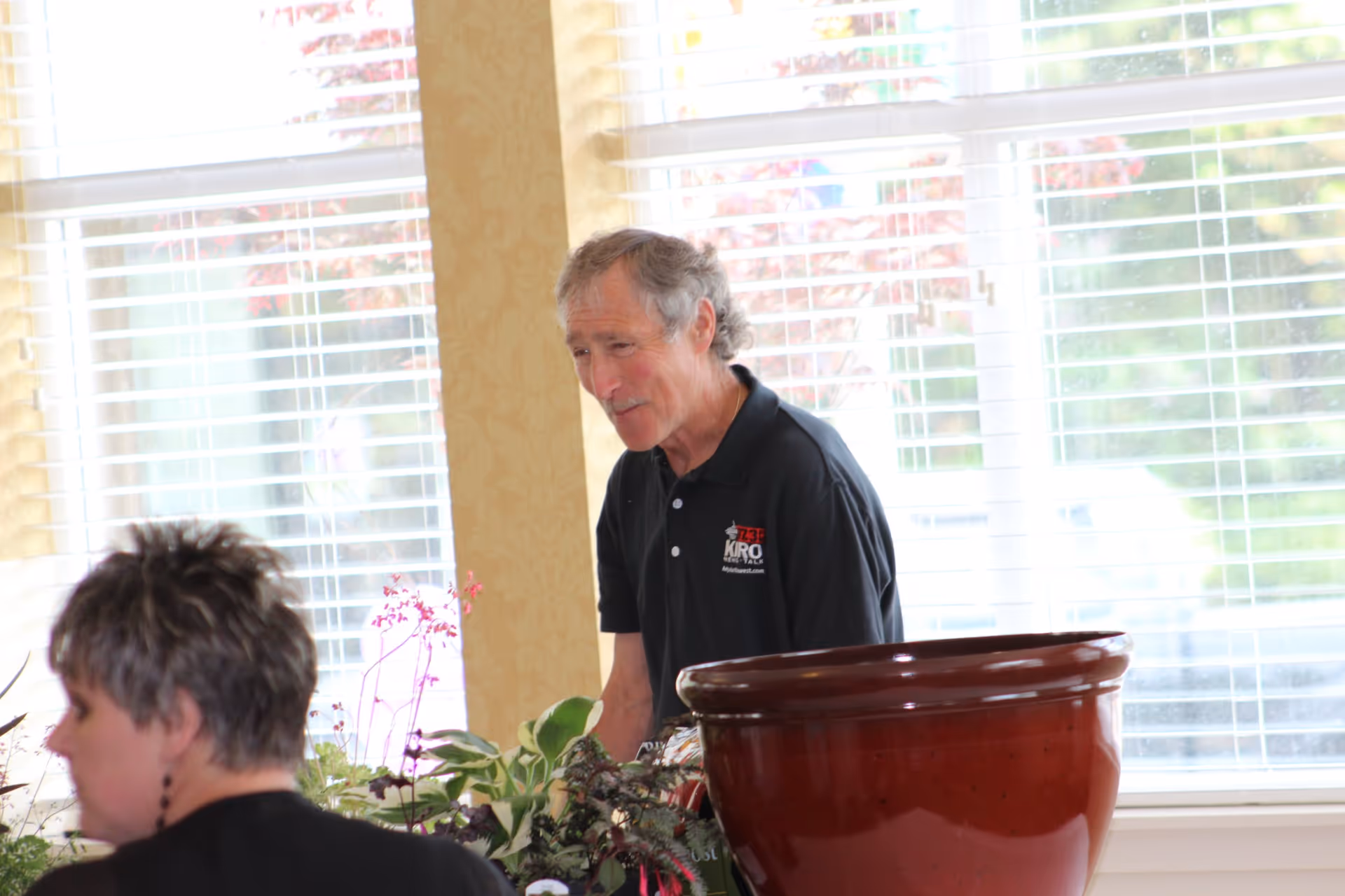 An older man wearing a black polo shirt with a logo is standing indoors near a large red ceramic pot and some plants, with a woman in the foreground and large windows with blinds in the background.