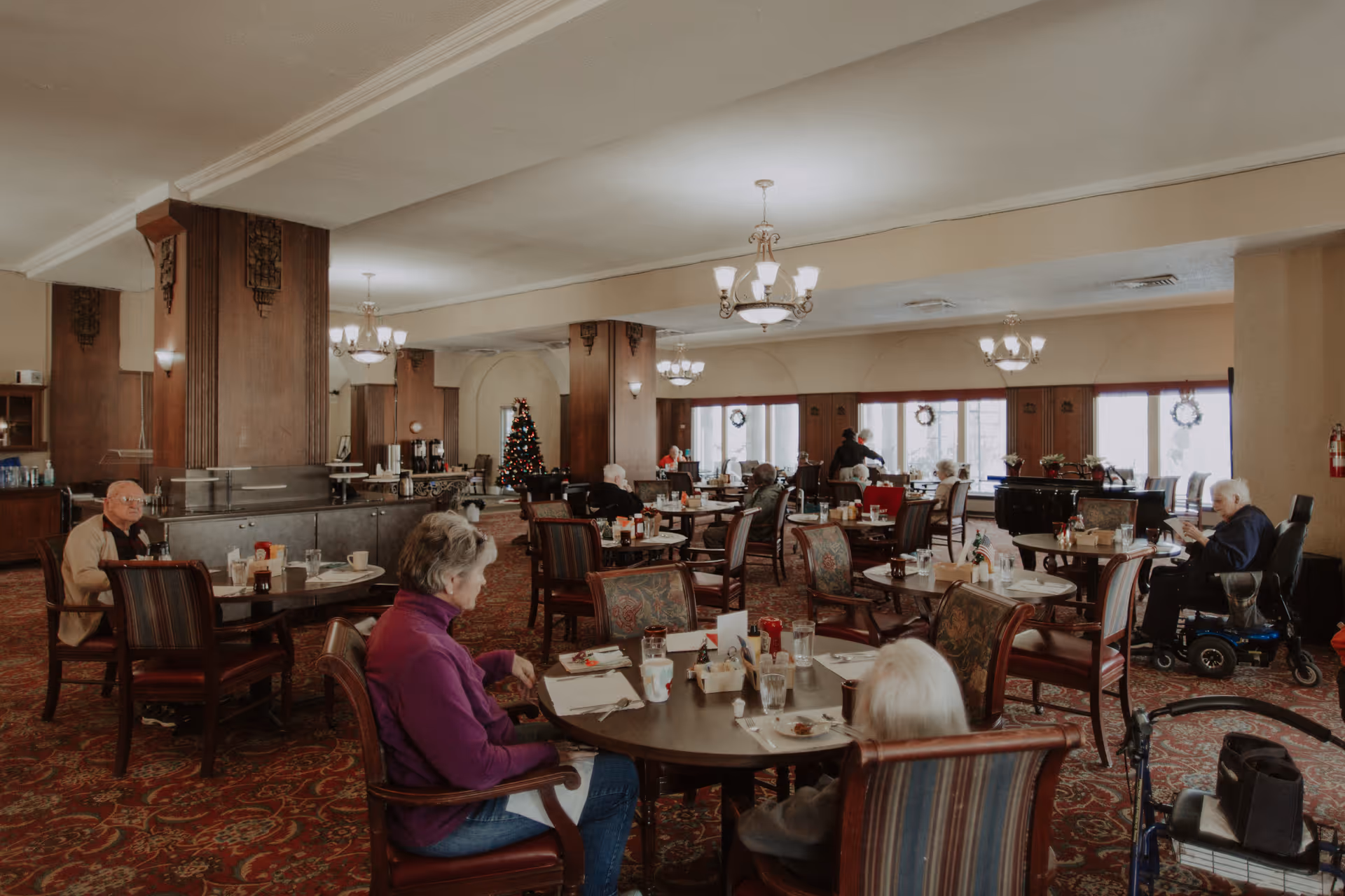 A spacious dining room in a senior living facility with several elderly residents seated at round tables. The room features patterned carpet, wooden pillars, chandeliers, and large windows with wreath decorations. A Christmas tree is visible in the background, and some residents are engaged in conversation or eating.