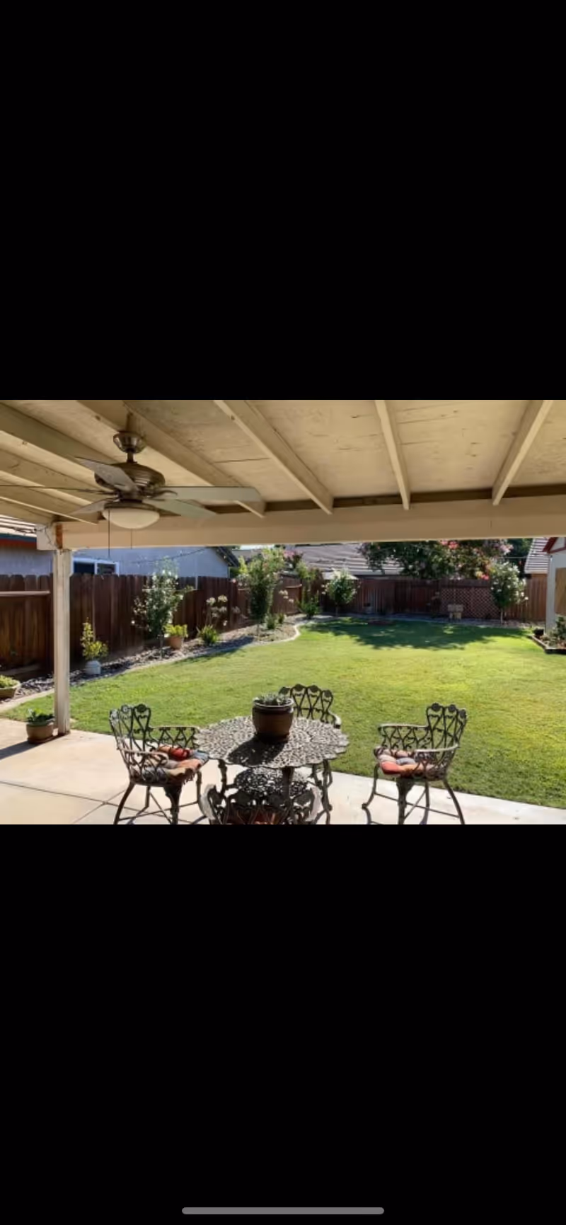 Covered patio area with a ceiling fan and a metal table with four chairs, each with a red cushion, overlooking a well-maintained backyard with green grass, a wooden fence, and various plants and shrubs along the fence line.