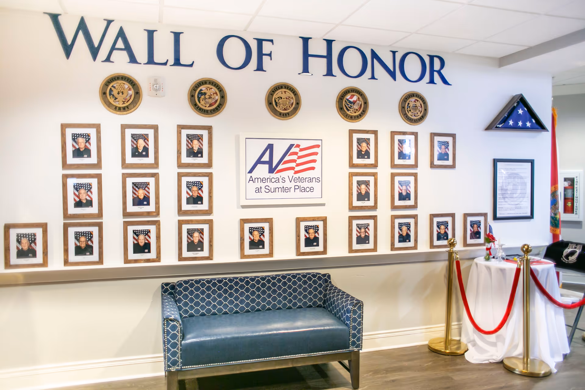 Interior wall display titled 'Wall of Honor' featuring framed photos of veterans with American flags in the background, military branch emblems above, and a folded American flag in a triangular case on the right. A blue patterned bench is placed below the display, and a small table with red ropes and a Florida state flag are visible to the right.