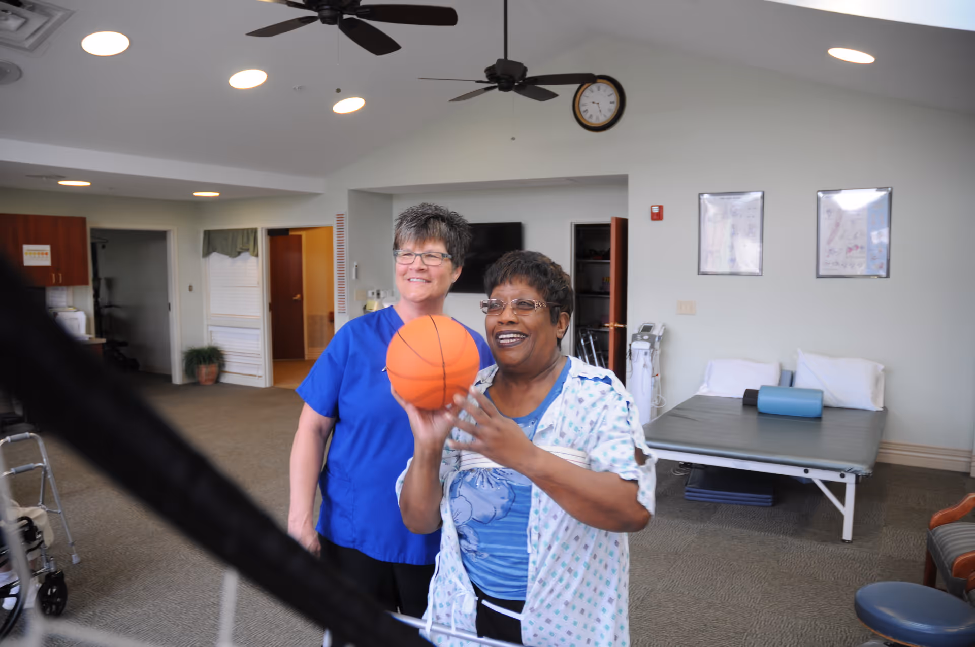 Two women in a therapy or activity room, one holding a small basketball and smiling, the other standing behind her also smiling. The room has a therapy bed with pillows and a bolster, medical equipment, ceiling fans, and framed posters on the wall.