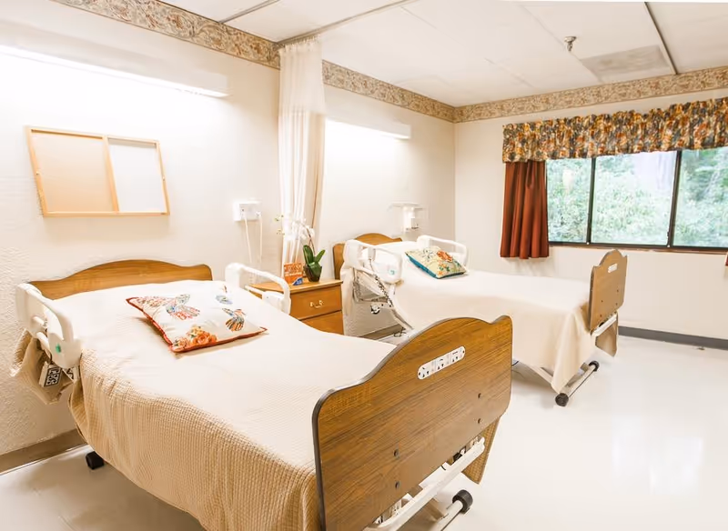 A bright room with two hospital-style beds, each with a floral pillow and beige bedspread. Between the beds is a wooden nightstand with a small plant and a decorative item. The room has a large window with floral curtains and a white ceiling with fluorescent lighting. The walls are light-colored with a decorative border near the ceiling.