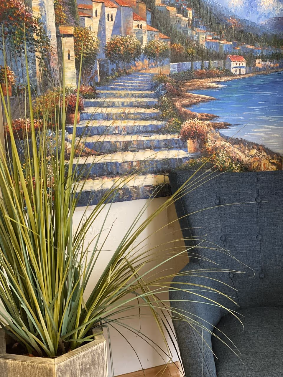 Potted tall grass plant next to a blue upholstered chair beneath a seaside painting on an interior wall.