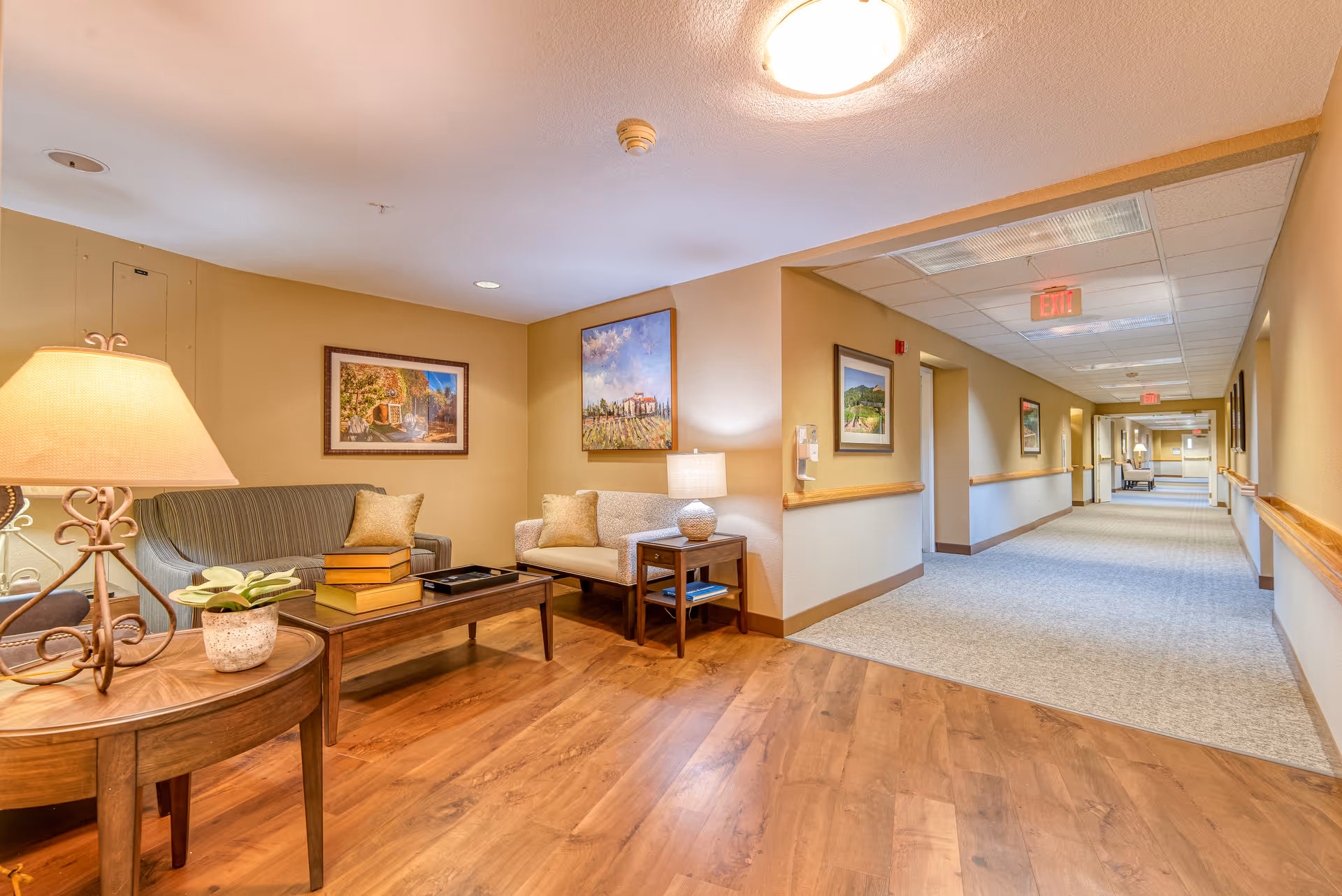 A well-lit senior living facility hallway with a seating area on the left side featuring a green sofa, a beige loveseat, a wooden coffee table with books and a tray, and a side table with a lamp and a small plant. The walls are decorated with framed paintings, and the floor transitions from wood to carpet as the hallway extends into the distance.