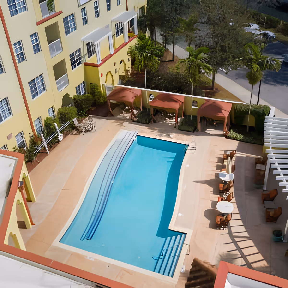 Aerial view of an outdoor swimming pool area at Discovery Village At Melbourne, surrounded by a yellow multi-story building with balconies, shaded cabanas, palm trees, and outdoor seating with tables and chairs.