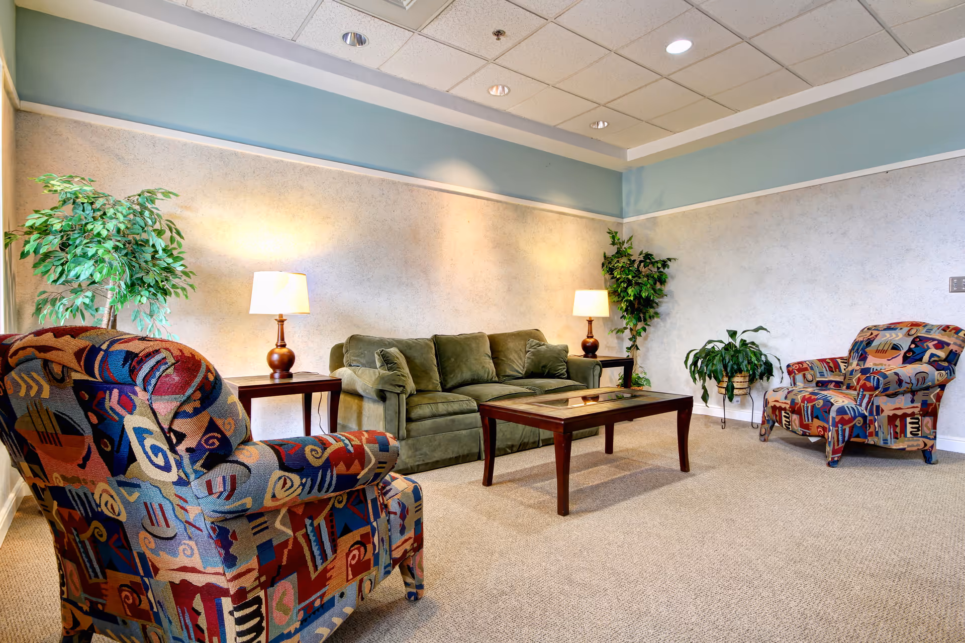 Comfortable common room with patterned armchairs, a green sofa, wooden coffee table, lamps, and potted plants.