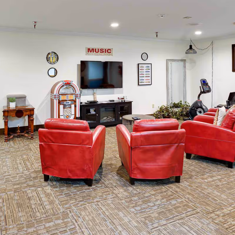 Communal living room with red leather armchairs facing a wall-mounted TV, jukebox, and small tables.