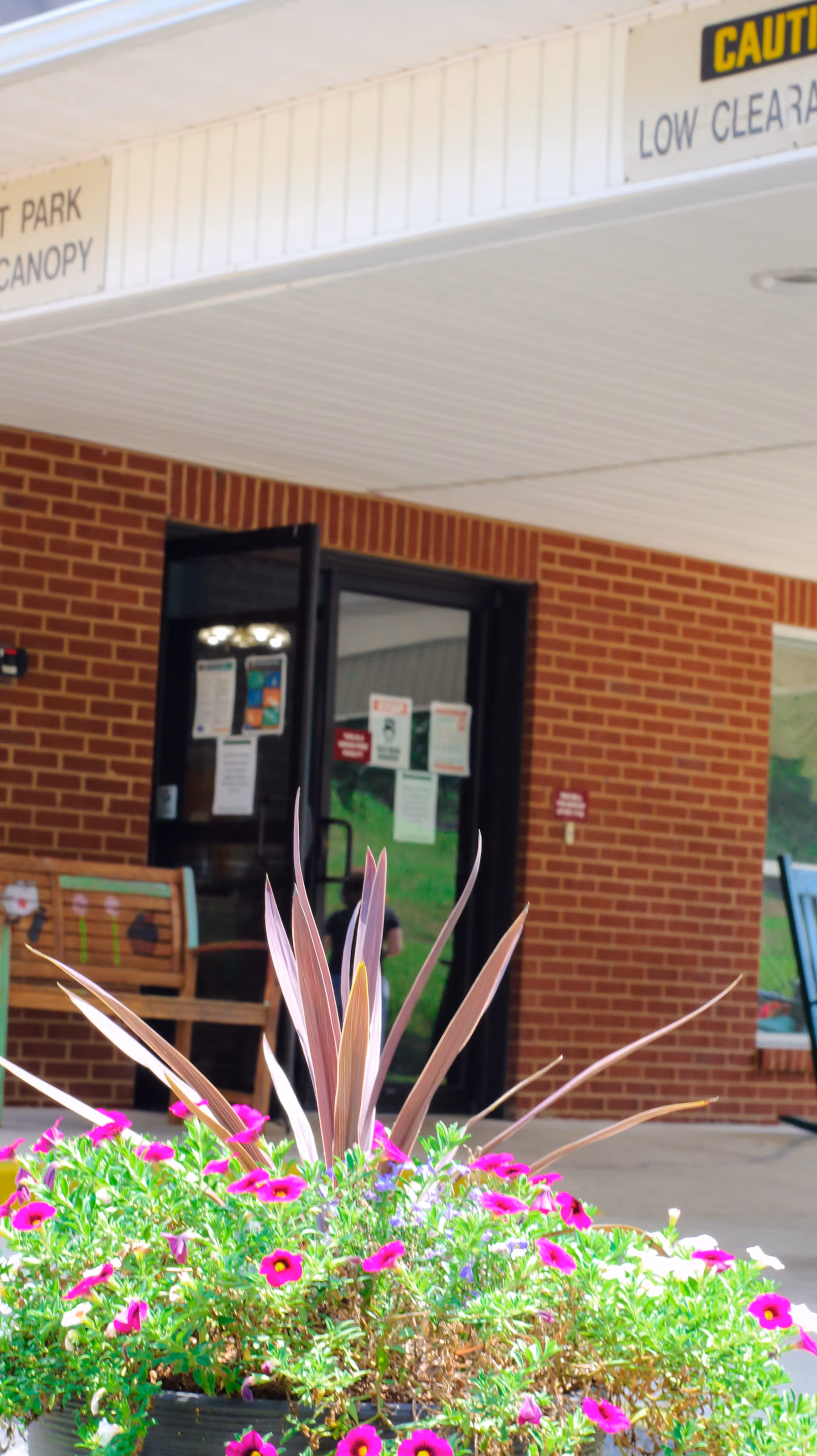Entrance area of Carriage Hill Retirement facility showing a brick wall with a glass door, a wooden bench with painted decorations, and a large planter filled with vibrant pink and purple flowers in the foreground. A white canopy overhead has caution signs about low clearance and parking.