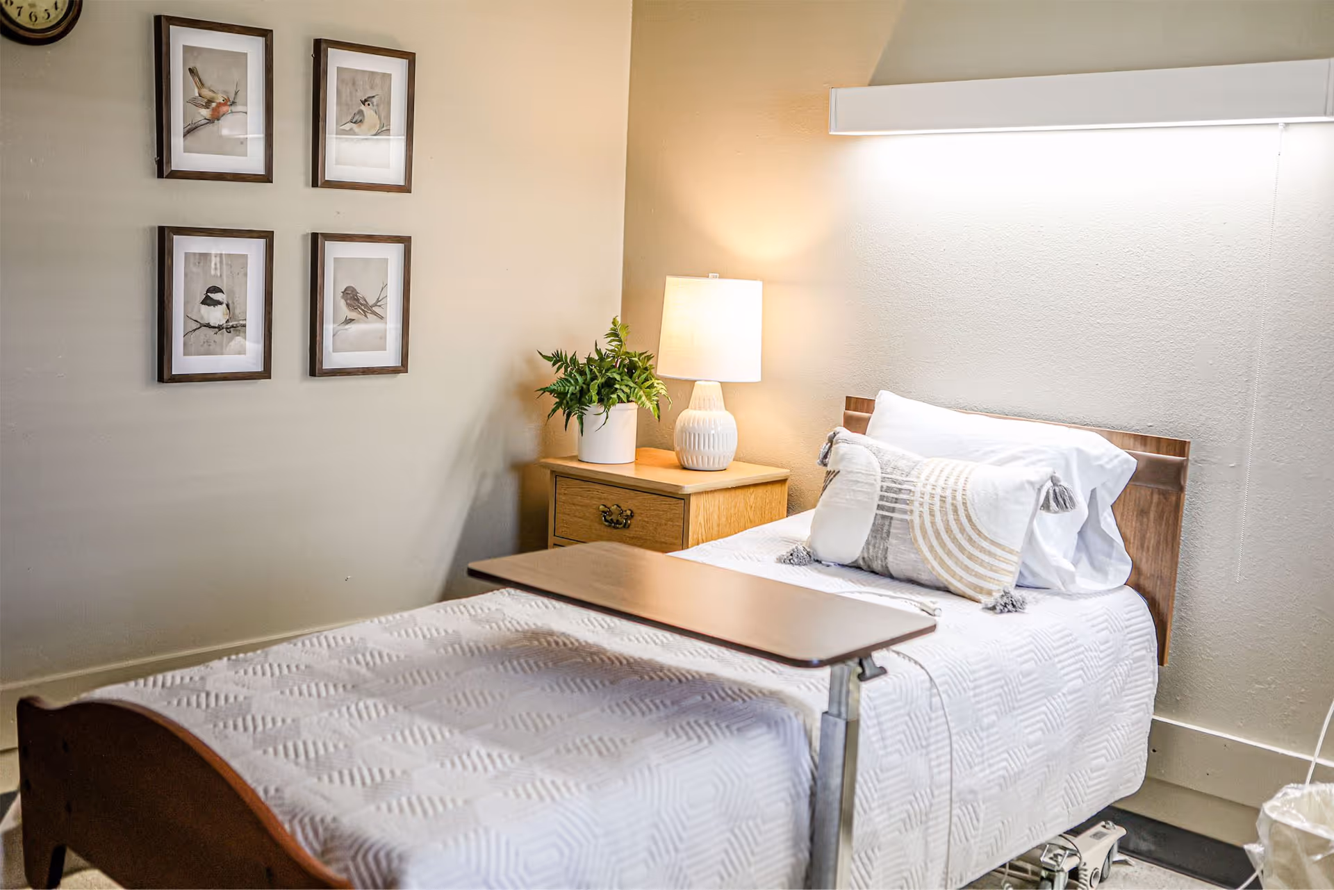 A neatly made single bed with white bedding and decorative pillows in a room. Next to the bed is a wooden nightstand with a white lamp and a potted plant. Four framed bird illustrations hang on the wall above the nightstand. A clock is partially visible on the wall. There is a wooden overbed table positioned over the bed.
