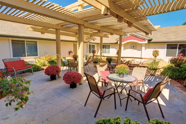 Outdoor patio area at Overland Court Senior Living with multiple seating arrangements including chairs and tables under a wooden pergola. The space is decorated with potted plants and flowers, surrounded by the building's exterior walls and windows.