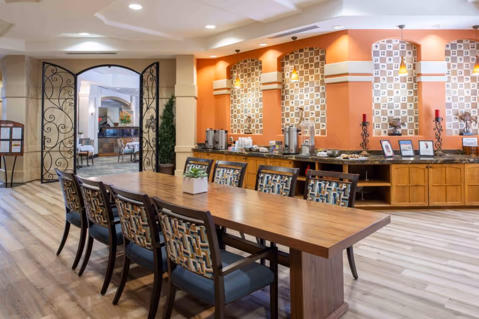 A spacious dining area with a long wooden table surrounded by eight chairs with patterned upholstery. The back wall features an orange accent with decorative tile insets and three pendant lights. A counter along the wall holds coffee urns, cups, and snacks. Decorative wrought iron double doors open to another room with additional seating and a fireplace.
