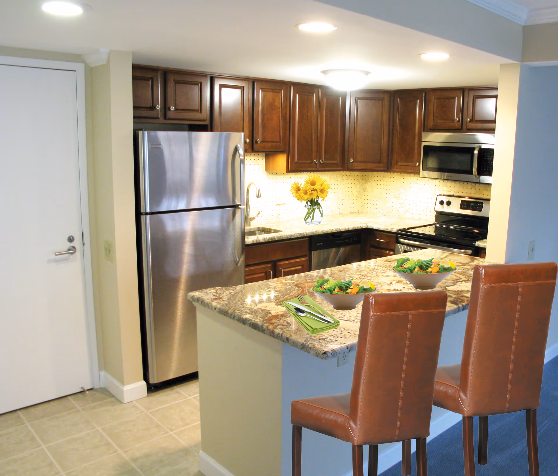 Modern kitchen with stainless steel refrigerator, stove, microwave, and dishwasher. Dark wooden cabinets and granite countertops with two brown leather bar stools at the counter. Two bowls of salad and a vase with yellow flowers on the counter.