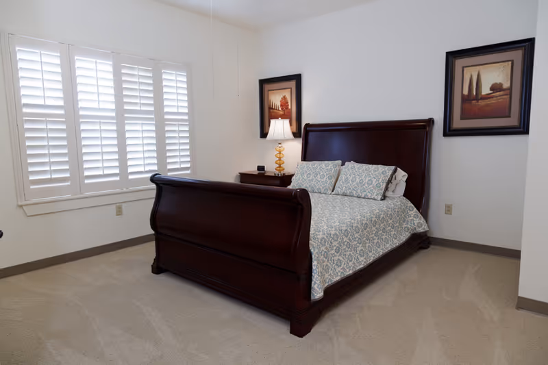 A bedroom with a dark wooden sleigh bed featuring a patterned bedspread and two matching pillows. There is a nightstand with a lamp and an alarm clock next to the bed. The room has beige carpet, white walls, two framed landscape paintings, and a window with white plantation shutters.