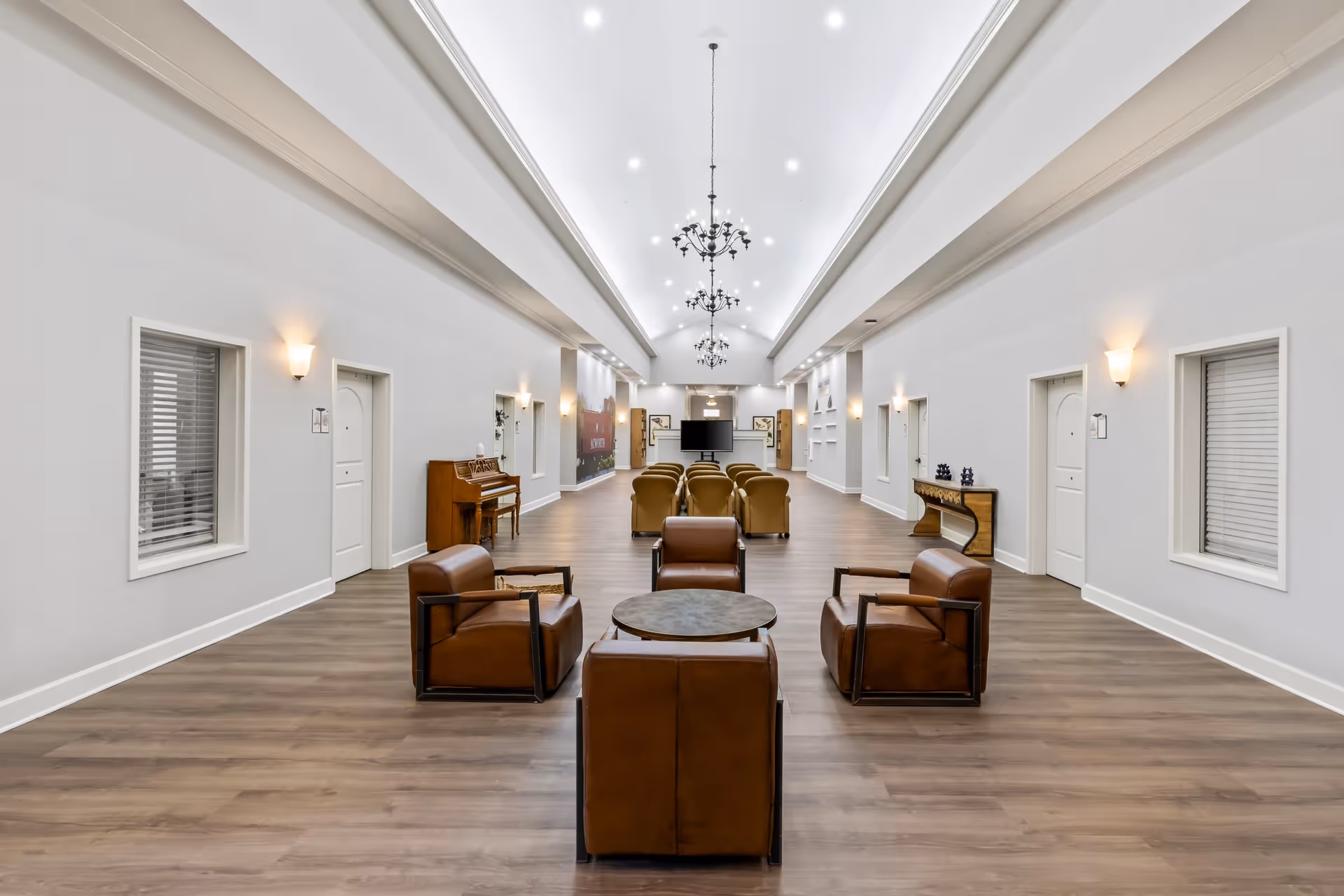 A spacious, well-lit common area with high ceilings and modern chandeliers. The room features a seating arrangement with four brown leather chairs around a small round table in the foreground and additional seating with armchairs facing a large flat-screen TV in the background. The walls are light gray with white trim, and there are several doors and windows along the sides. A piano and a console table with decorative items are also visible.
