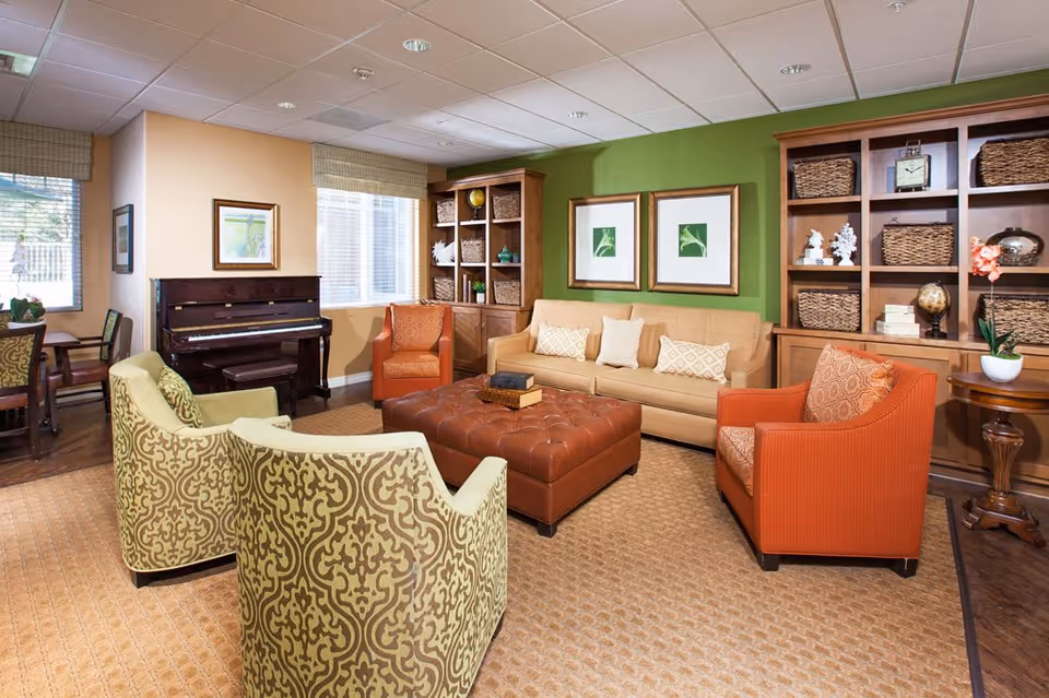 A cozy living room with a beige sofa, two orange armchairs, and two patterned green armchairs arranged around a large brown tufted ottoman. Behind the sofa is a green accent wall with two framed botanical prints. On either side of the sofa are wooden bookshelves filled with wicker baskets and decorative items. A piano is positioned near two windows with beige blinds, and a small round wooden table with a potted orchid is on the right side.