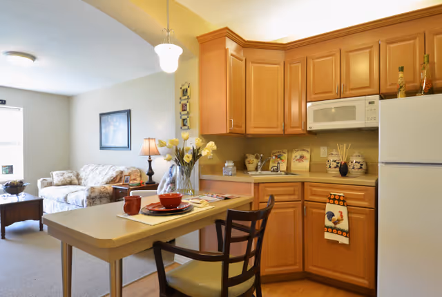A cozy kitchen and living room area in a senior living facility. The kitchen features wooden cabinets, a white microwave, and a refrigerator. A small dining counter with two chairs is set with red dishes and a vase of flowers. The living room has a floral patterned sofa, a wooden coffee table, a side table with a lamp, and a framed picture on the wall.