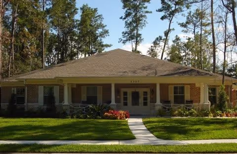 Single-story brick building with a wide front porch, surrounded by a well-maintained lawn and tall trees in the background under a clear sky.