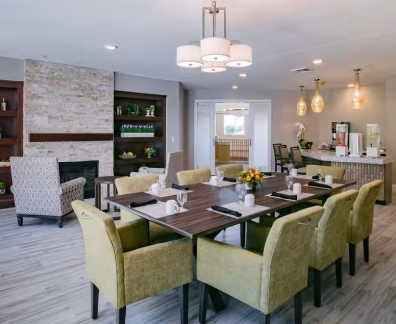 A modern dining room with a rectangular wooden table set for eight people with green upholstered chairs. The table has white placemats, black napkins, white mugs, and glasses. In the background, there is a stone fireplace with built-in shelves containing decorative items, two patterned armchairs, and a counter area with beverage dispensers and three hanging pendant lights. The room has light-colored flooring and recessed ceiling lights.