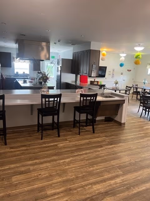 Interior view of a communal dining and kitchen area in a senior living facility. The foreground shows a counter with three black chairs and name tags on the counter. The kitchen area in the background has dark cabinets, a stainless steel range hood, and various kitchen appliances. The dining area has several tables and chairs, with colorful hanging decorations from the ceiling.