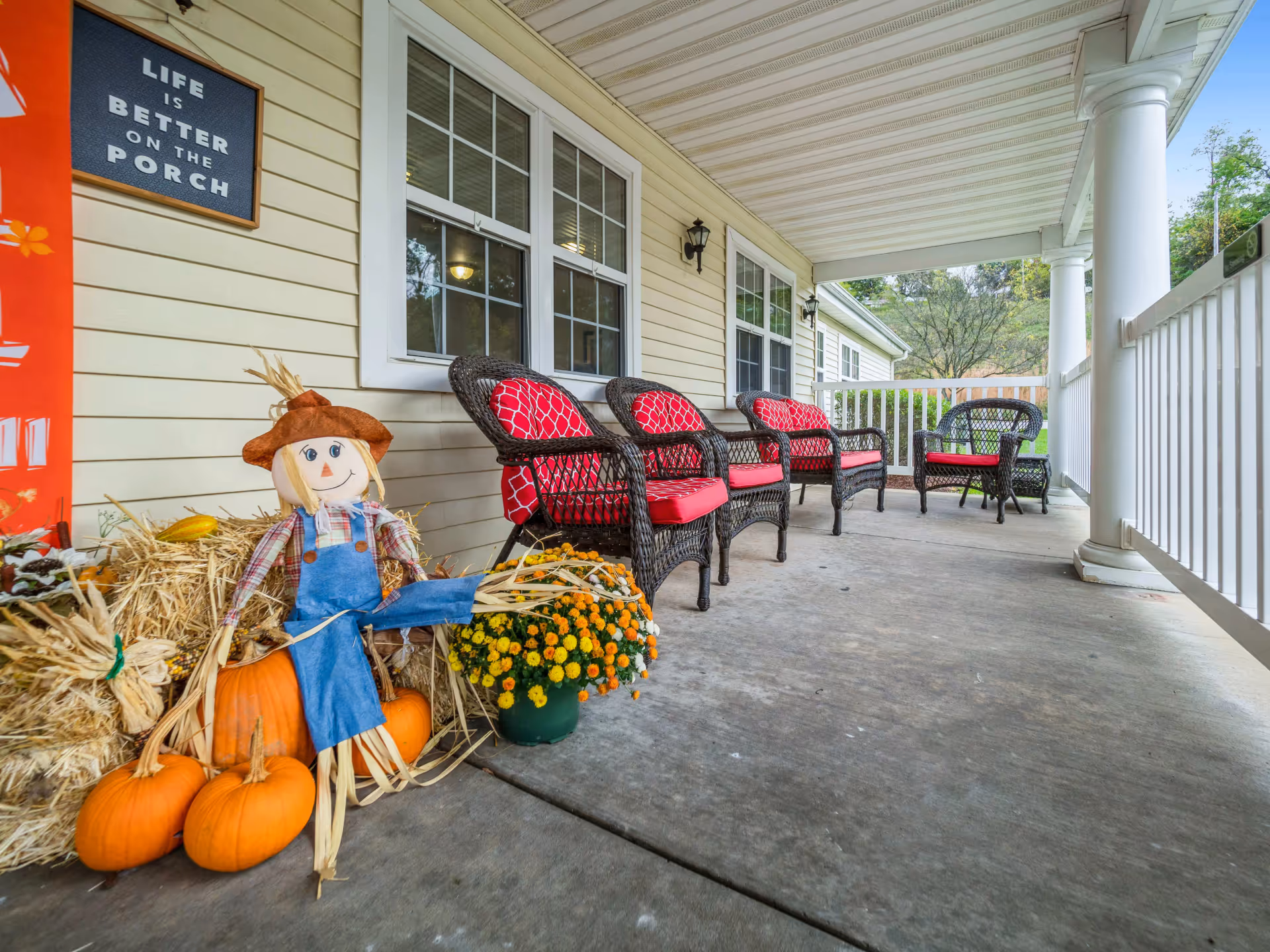 Covered porch area with wicker chairs featuring red cushions lined up against the wall of a building. Seasonal decorations including a scarecrow, pumpkins, hay bales, and a pot of yellow and orange flowers are arranged near the entrance. A sign on the wall reads 'Life is better on the porch.'