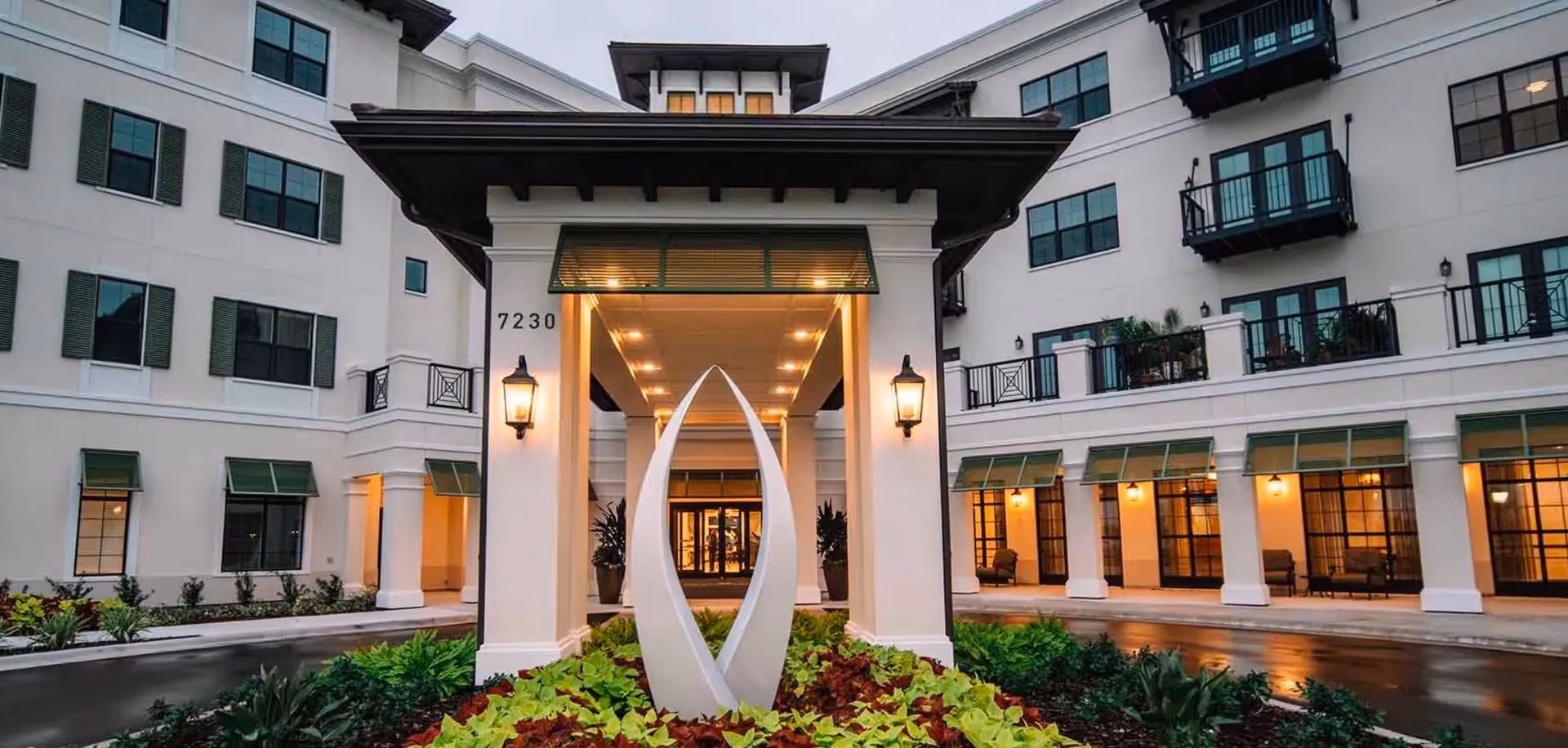 Entrance of a multi-story senior living facility with a covered driveway, modern sculpture in the center of a landscaped garden, and warm lighting illuminating the building exterior and windows.