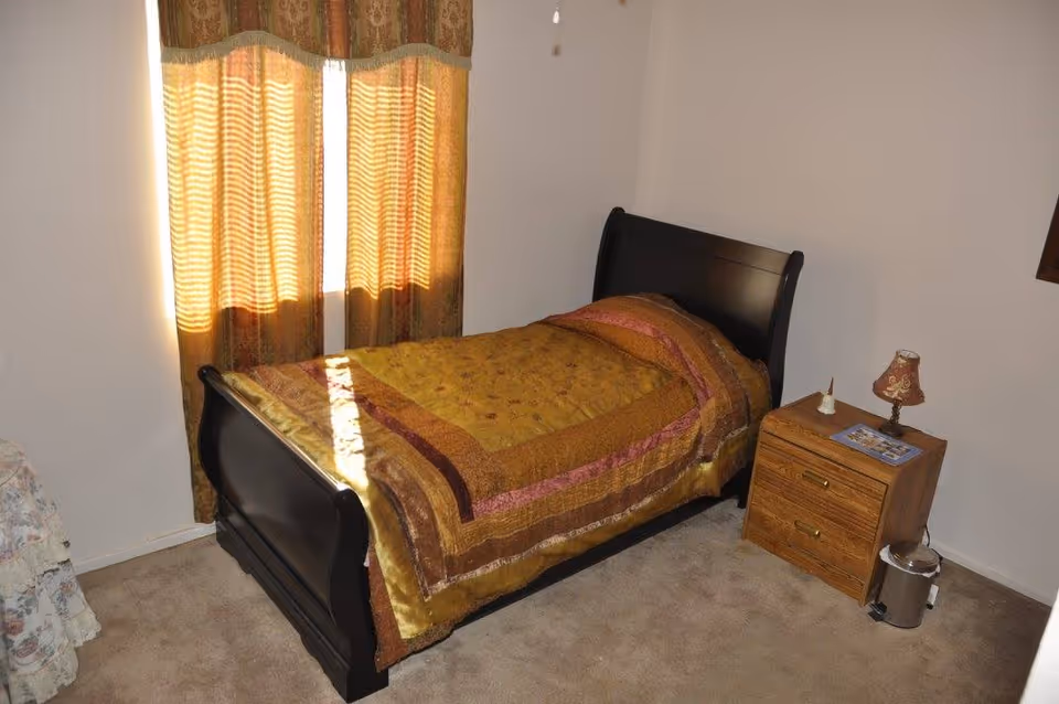 Single bed with a patterned bedspread next to a nightstand and curtained window in a small bedroom.