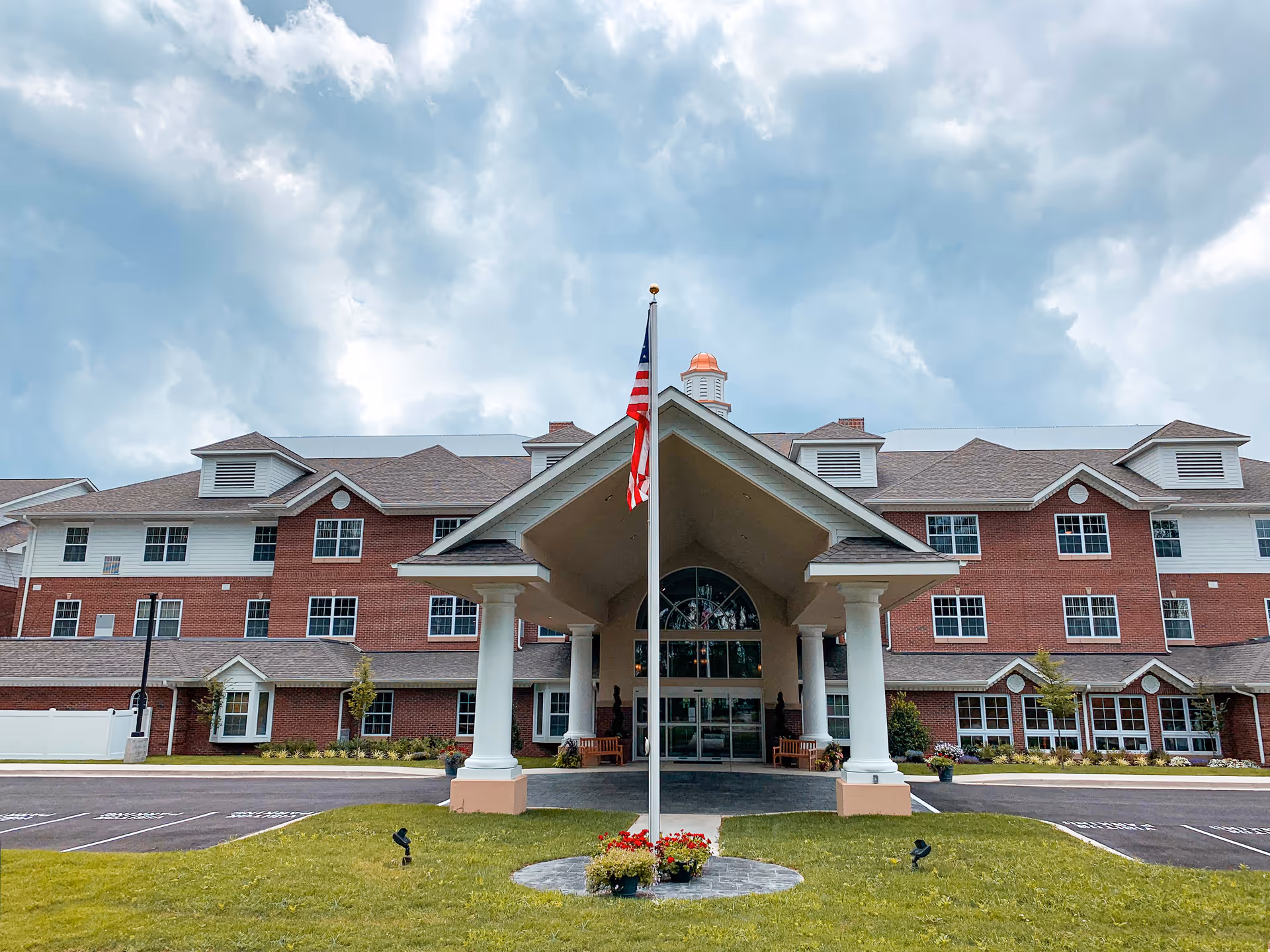 Front exterior view of South Hills Square Retirement Resort, a large three-story building with red brick and white siding, featuring a covered entrance supported by white columns and an American flag on a flagpole in front. The sky is partly cloudy.