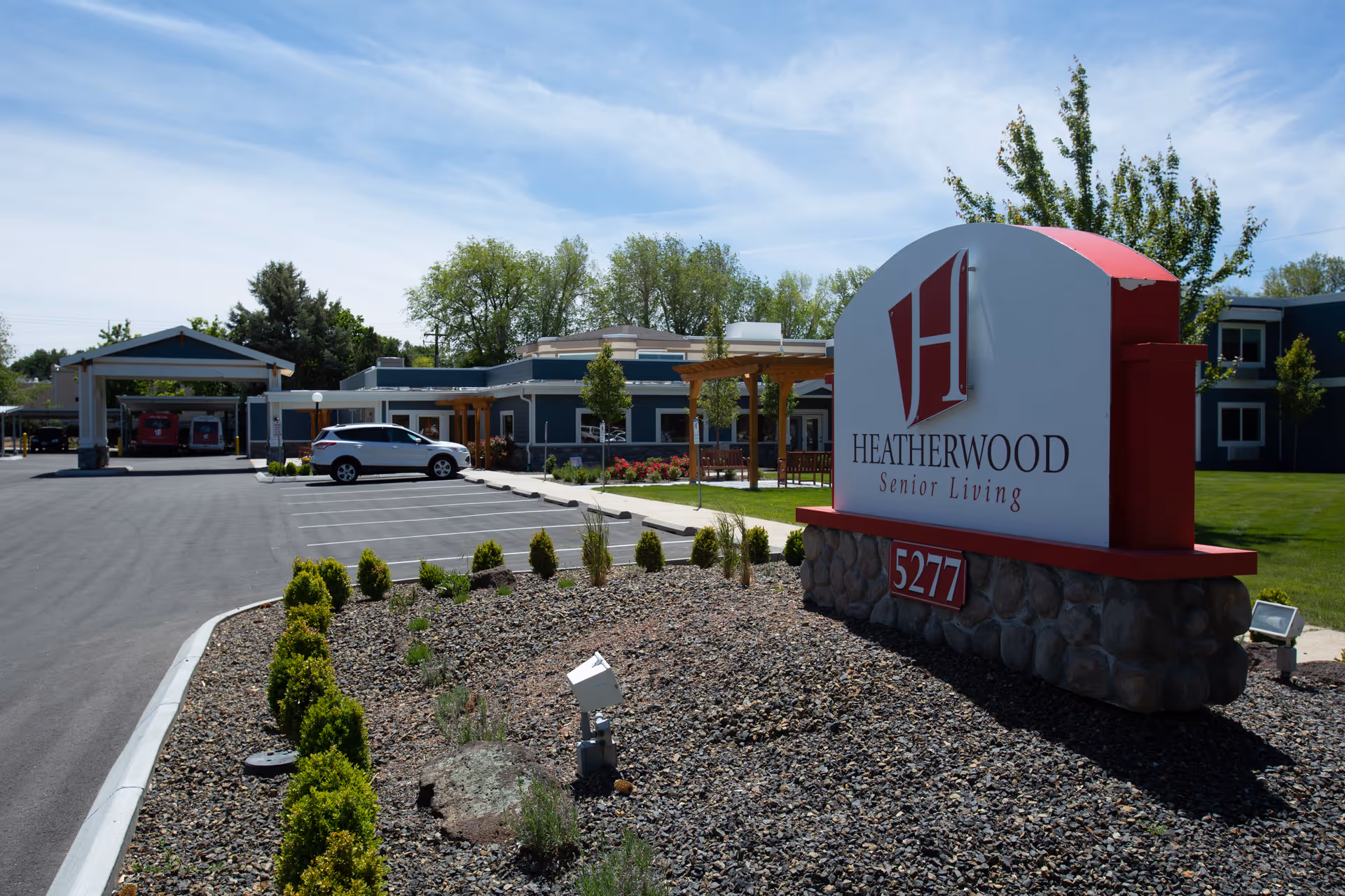 Exterior view of Heatherwood Senior Living facility showing the main entrance sign with the address 5277, a parking lot with a white SUV, landscaped area with small bushes and rocks, and the building in the background under a partly cloudy sky.