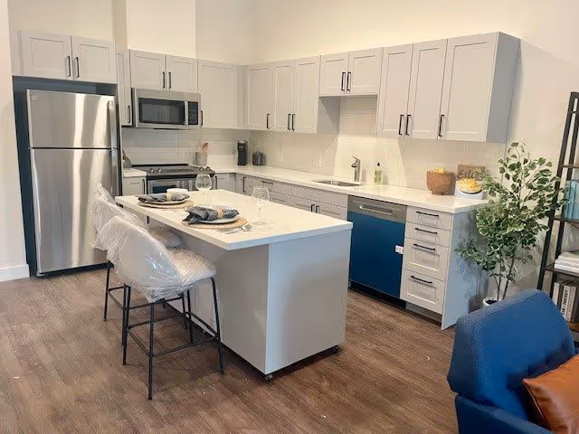 Modern kitchen with white cabinets, stainless steel refrigerator, microwave, stove, and dishwasher. A kitchen island with two bar stools covered in plastic is set with plates and glasses. The floor is wooden, and there is a potted plant and a blue chair visible on the right side.
