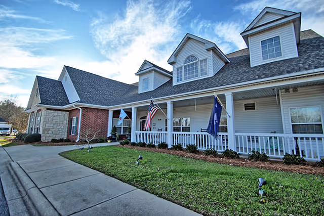 Exterior view of Sumter Terrace Assisted Living facility showing a large building with a porch, white railing, and multiple dormer windows on the roof. There are three flags displayed on the porch, and a well-maintained lawn and sidewalk in front of the building under a partly cloudy sky.