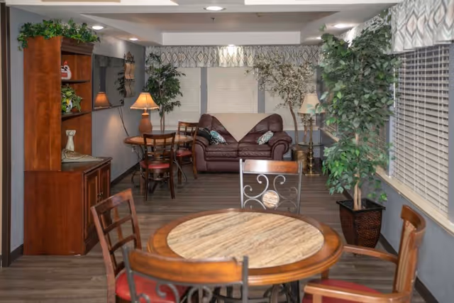 A cozy common area with a round wooden table and chairs in the foreground, a brown leather couch with pillows and a throw blanket against the back wall, several potted plants, a wooden cabinet with decorative items, a wall-mounted TV, and table lamps providing warm lighting.