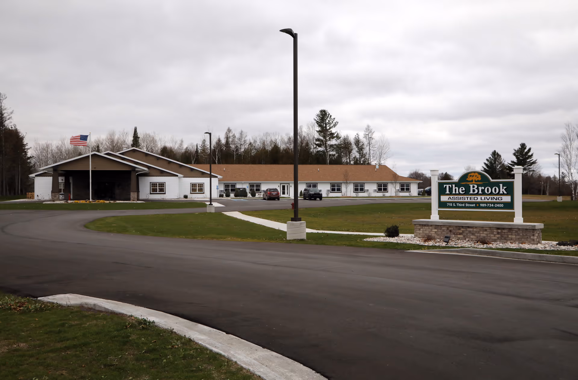 Exterior view of The Brook Assisted Living facility in Rogers City, showing a single-story building with a brown roof and white walls, an American flag on a flagpole, a paved driveway, and a sign with the facility's name and contact information on a grassy lawn under a cloudy sky.