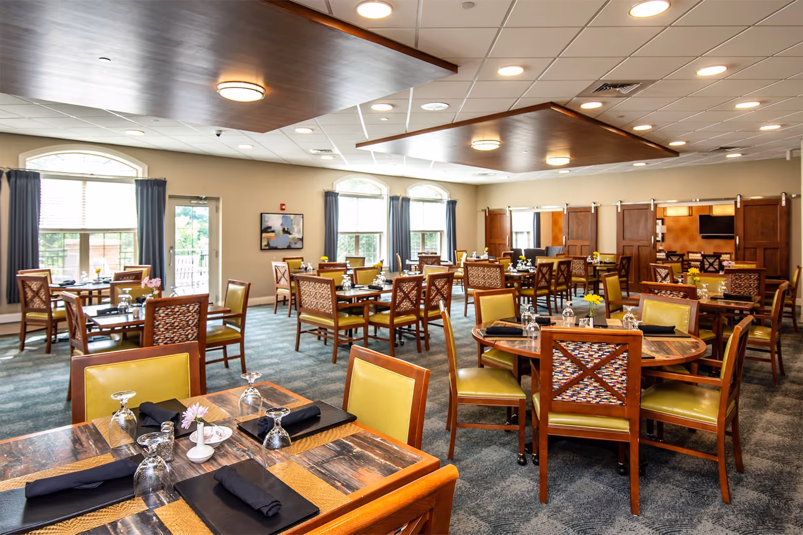 Bright, spacious dining room with multiple round and square tables set for meals and wooden chairs.