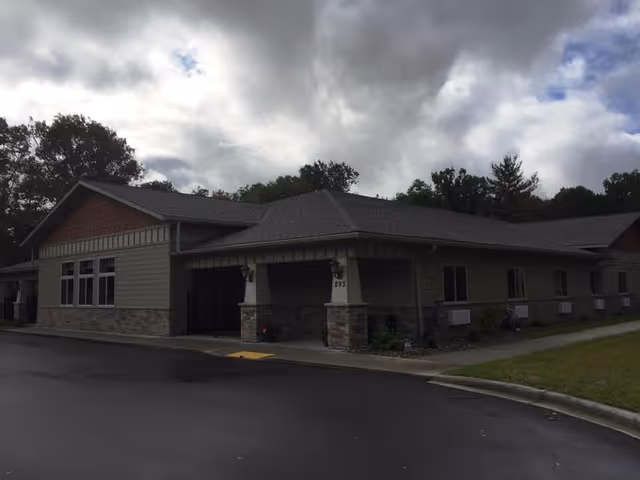 Exterior view of a single-story assisted living facility building with a covered entrance, surrounded by trees and a cloudy sky overhead.