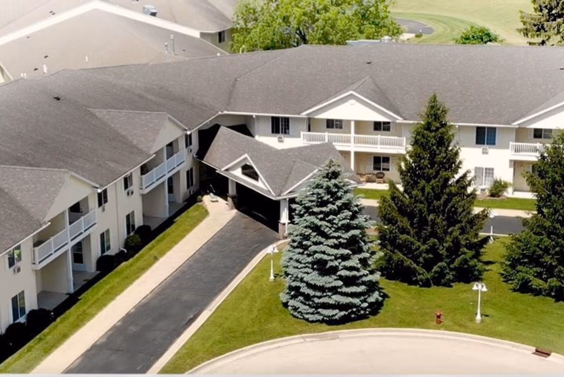 Aerial view of Charleston House Assisted Living facility showing a two-story building with a gray roof, white exterior walls, balconies, and a covered entrance driveway. The surrounding area includes green lawns, several large evergreen trees, and a curved driveway.