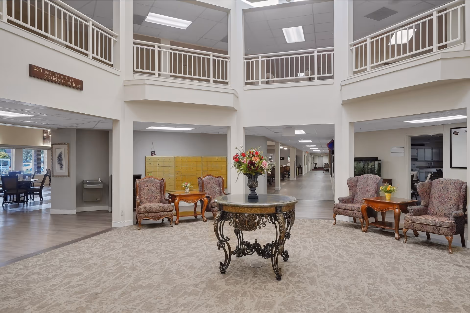 A spacious and well-lit common area in a senior living facility featuring a round ornate table with a floral arrangement in the center. Surrounding the table are several upholstered armchairs and small wooden side tables with flowers. The area has a high ceiling with a balcony railing above and multiple hallways leading to other parts of the facility. There is a sign on the wall that reads 'Don't just live with us, participate with us!'