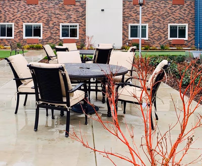 Outdoor courtyard with a round metal table and cushioned chairs on a wet concrete patio in front of a brick building.
