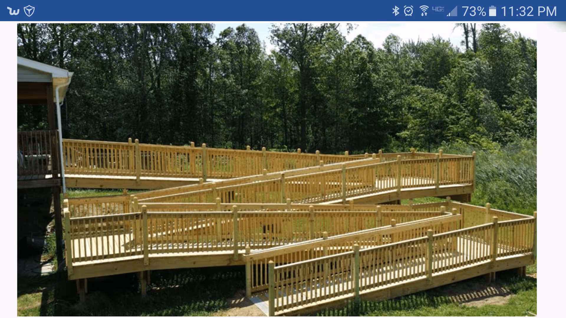 Outdoor wooden wheelchair ramp with multiple switchbacks leading up to a building, surrounded by green grass and trees in the background.