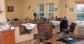 Interior of a hair salon area with two salon chairs, two hair washing sinks, a counter with hair care products, a basket with towels, and windows letting in natural light.
