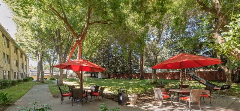Shaded outdoor courtyard with red patio umbrellas, tables and chairs beside a senior living building.